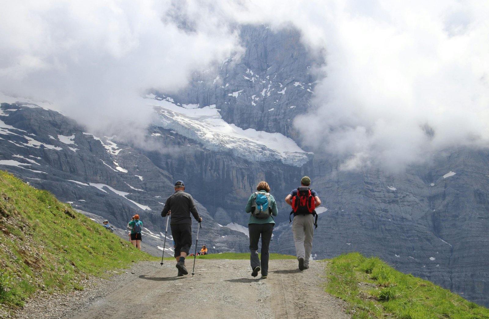 Group of hikers trekking on a mountain path with snow-capped peaks and cloudy skies.